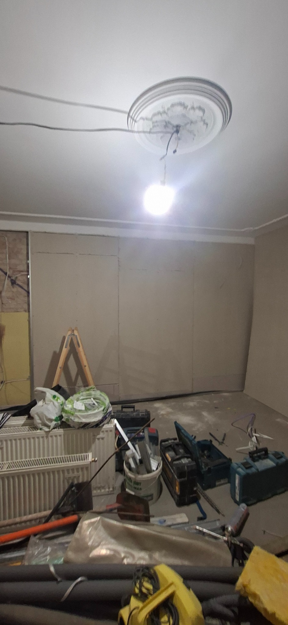 Pokój w trakcie remontu: ściany wyłożone płytami kartonowo-gipsowymi, widoczne narzędzia i materiały budowlane, drabina oparta o ścianę. Centralne oświetlenie sufitowe.