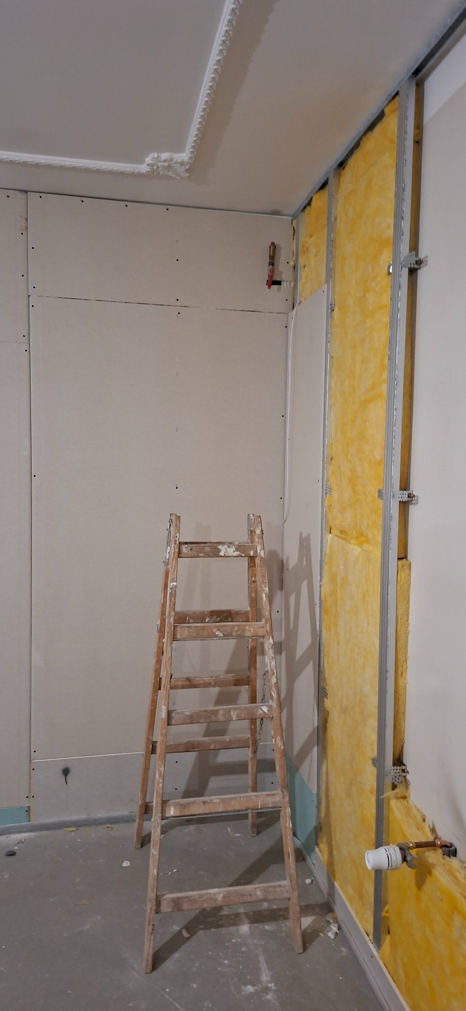 Ściana w trakcie wykańczania: płyty gipsowe na jednej ścianie, ocieplenie wełną mineralną na drugiej, drabina malarska w pomieszczeniu. Widoczne profile i instalacje.