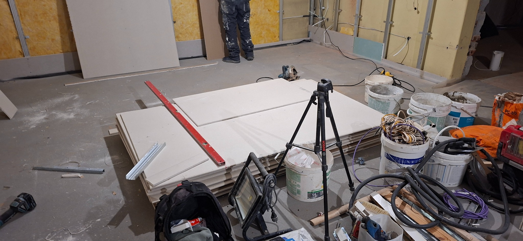 Stos płyt kartonowo-gipsowych w pomieszczeniu w trakcie prac wykończeniowych. Widoczne narzędzia, poziomica, oświetlenie robocze i częściowo ocieplona ściana.