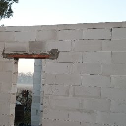 Matbud - Wnętrze domu w trakcie budowy z białych bloczków, widoczne nadproże okienne z cegieł. Na ścianie widoczne notatki. W tle zieleń i mgła.