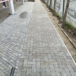 Matbud - Ułożona kostka brukowa w dwóch wzorach na zewnątrz budynku. Część kostki ułożona pod kątem, druga w klasyczny wzór. Widoczne zabezpieczenia budowy.