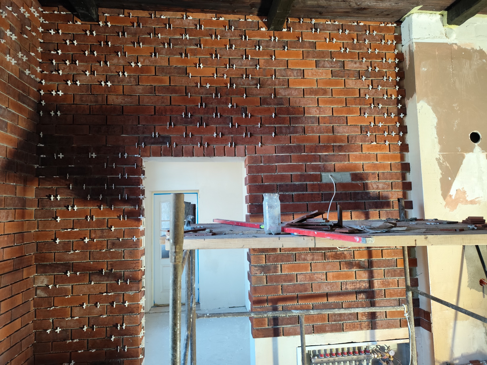 Układanie cegieł klinkierowych na ścianie z użyciem krzyżyków dystansowych. Widoczne rusztowanie i narzędzia. Częściowo wykończona ściana z cegły klinkierowej.