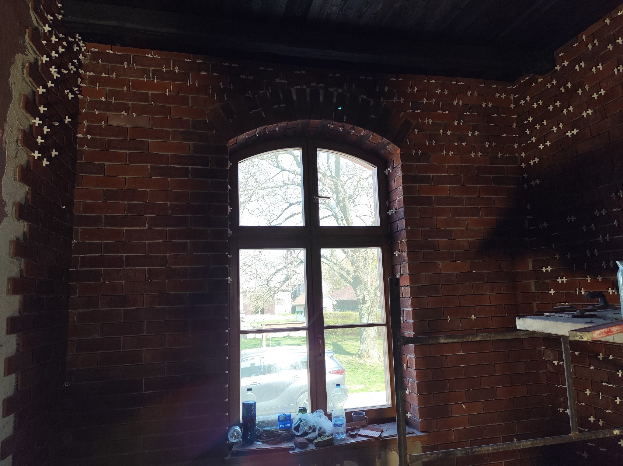 Układanie cegieł klinkierowych na ścianie wewnętrznej z oknem łukowym. Widoczne krzyżyki dystansowe. Częściowo wykończona ściana z cegły w odcieniach czerwieni i brązu.
