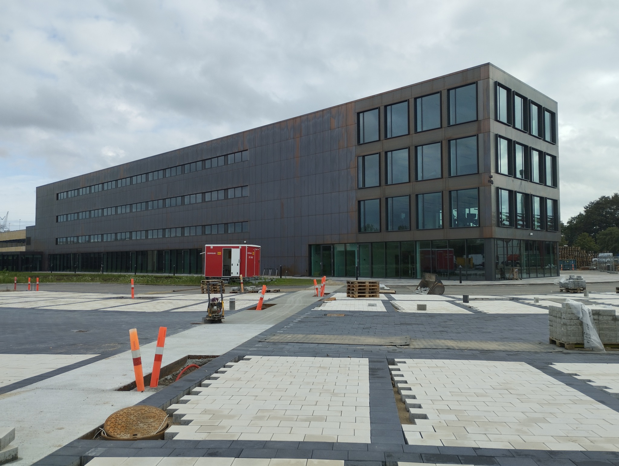 Układanie kostki brukowej na placu przed nowoczesnym budynkiem biurowym. Widoczne narzędzia i materiały budowlane. Prace w toku, częściowo ułożona kostka.