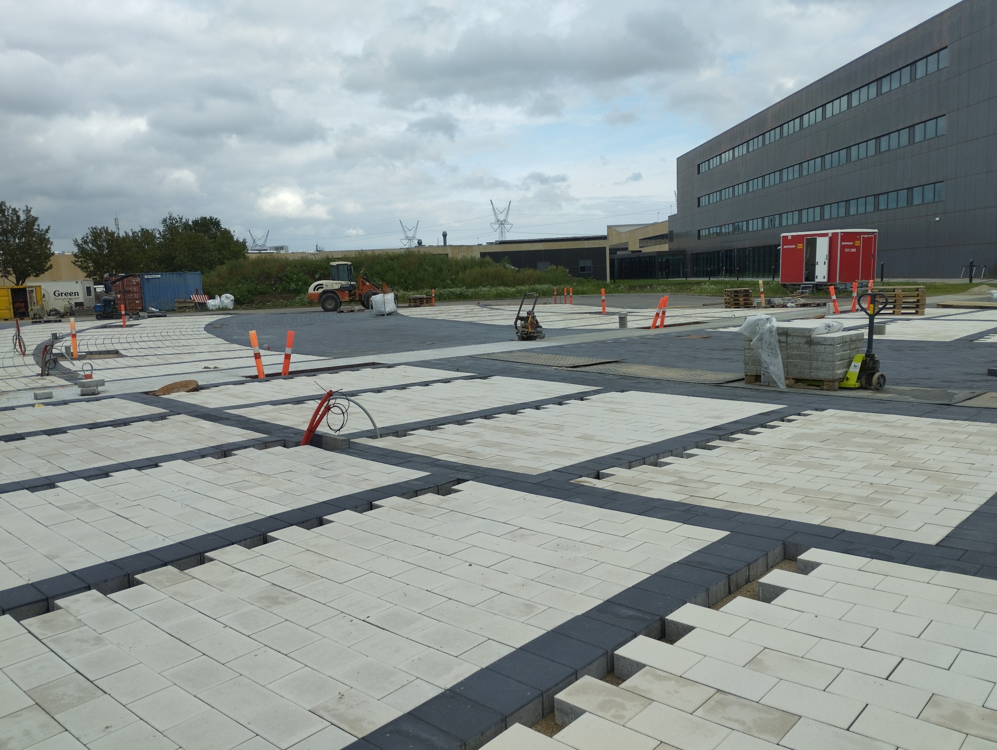 Układanie jasnoszarej kostki brukowej z ciemnymi obrzeżami na placu budowy. Widoczne maszyny budowlane i materiały. W tle budynek i pochmurne niebo.