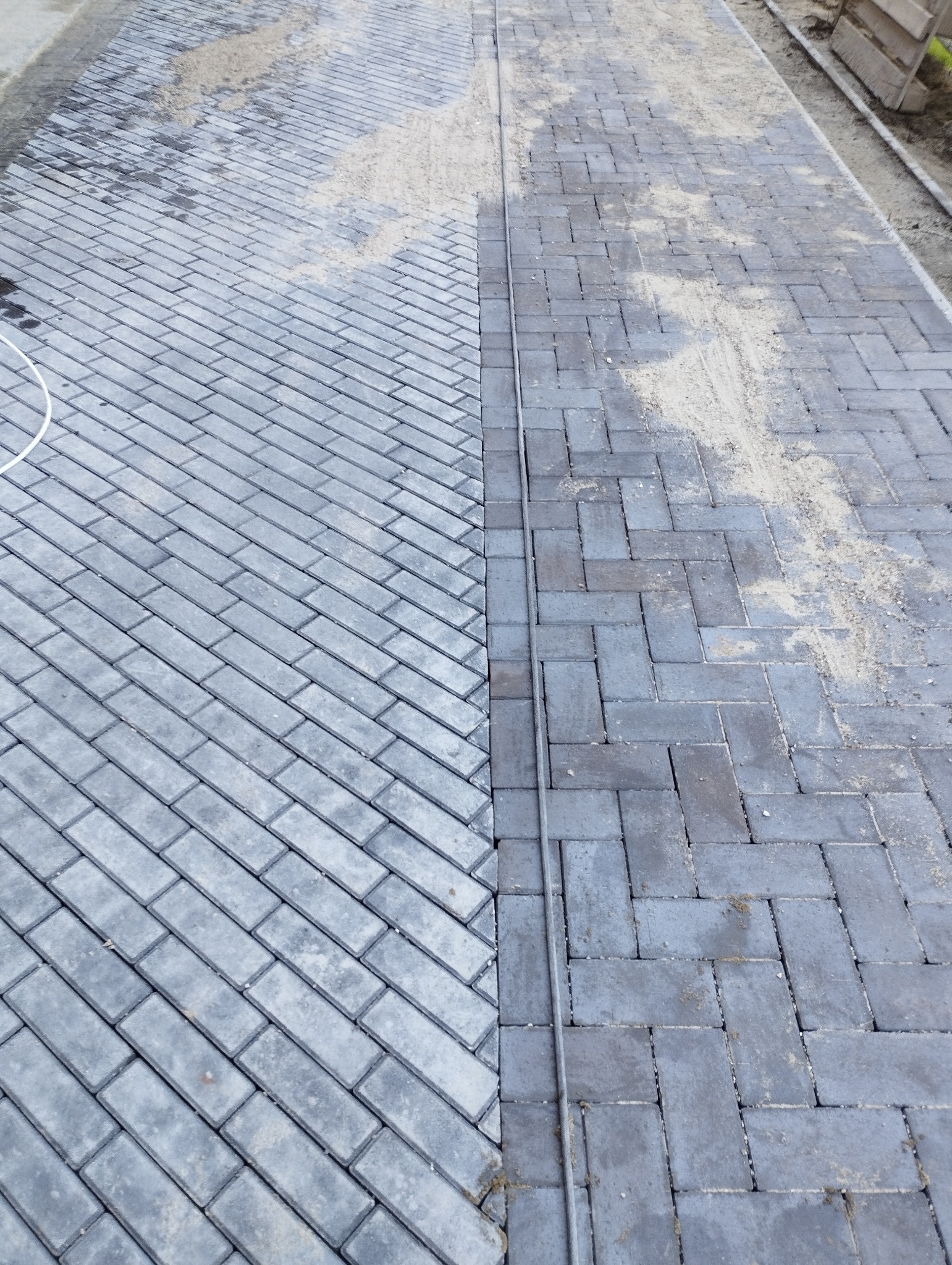 Ułożona kostka brukowa w dwóch odcieniach szarości, oddzielona linią. Część powierzchni posypana piaskiem, co sugeruje etap prac wykończeniowych.