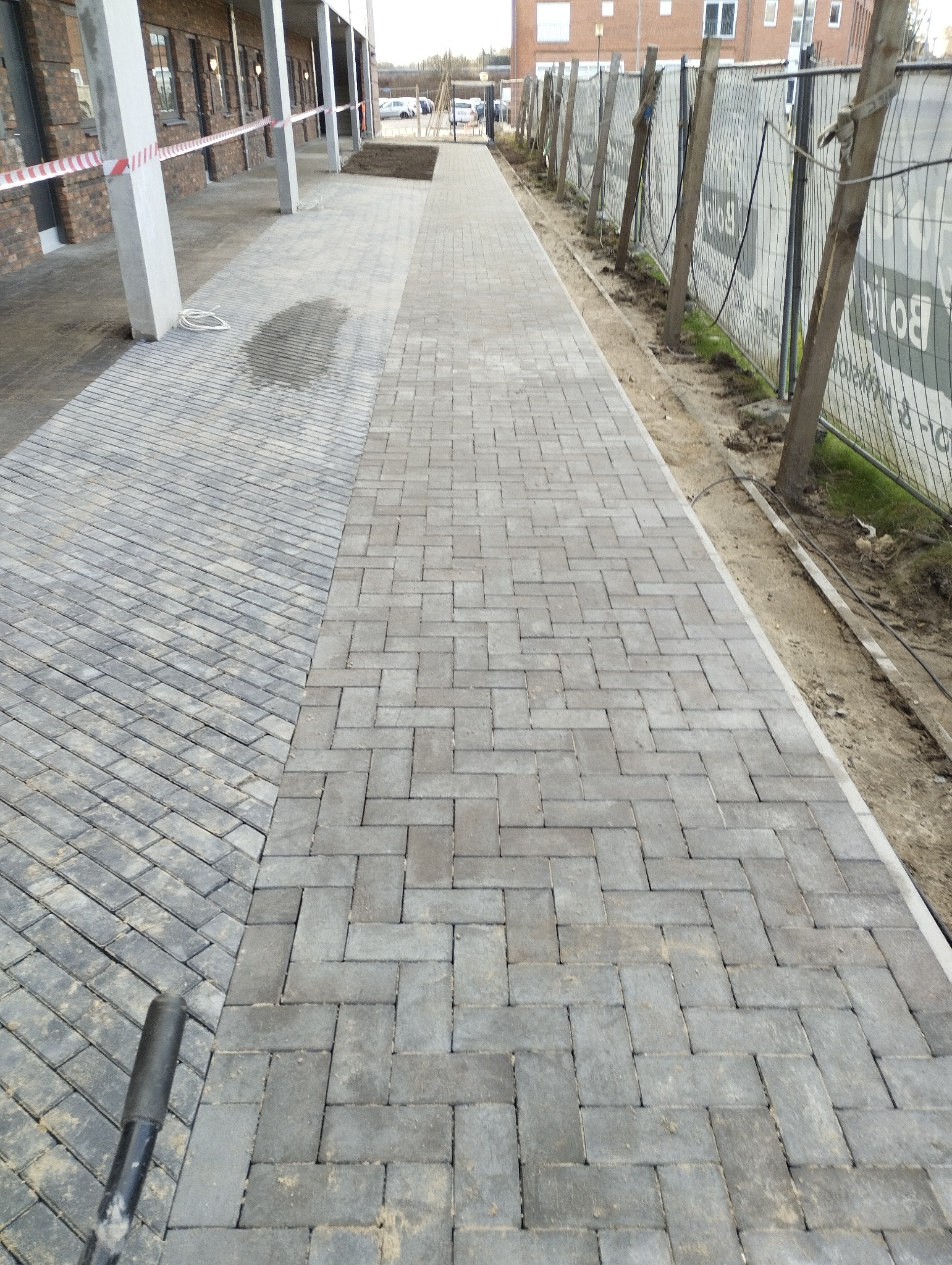 Ułożona kostka brukowa w dwóch wzorach na zewnątrz budynku. Część kostki ułożona pod kątem, druga w klasyczny wzór. Widoczne zabezpieczenia budowy.