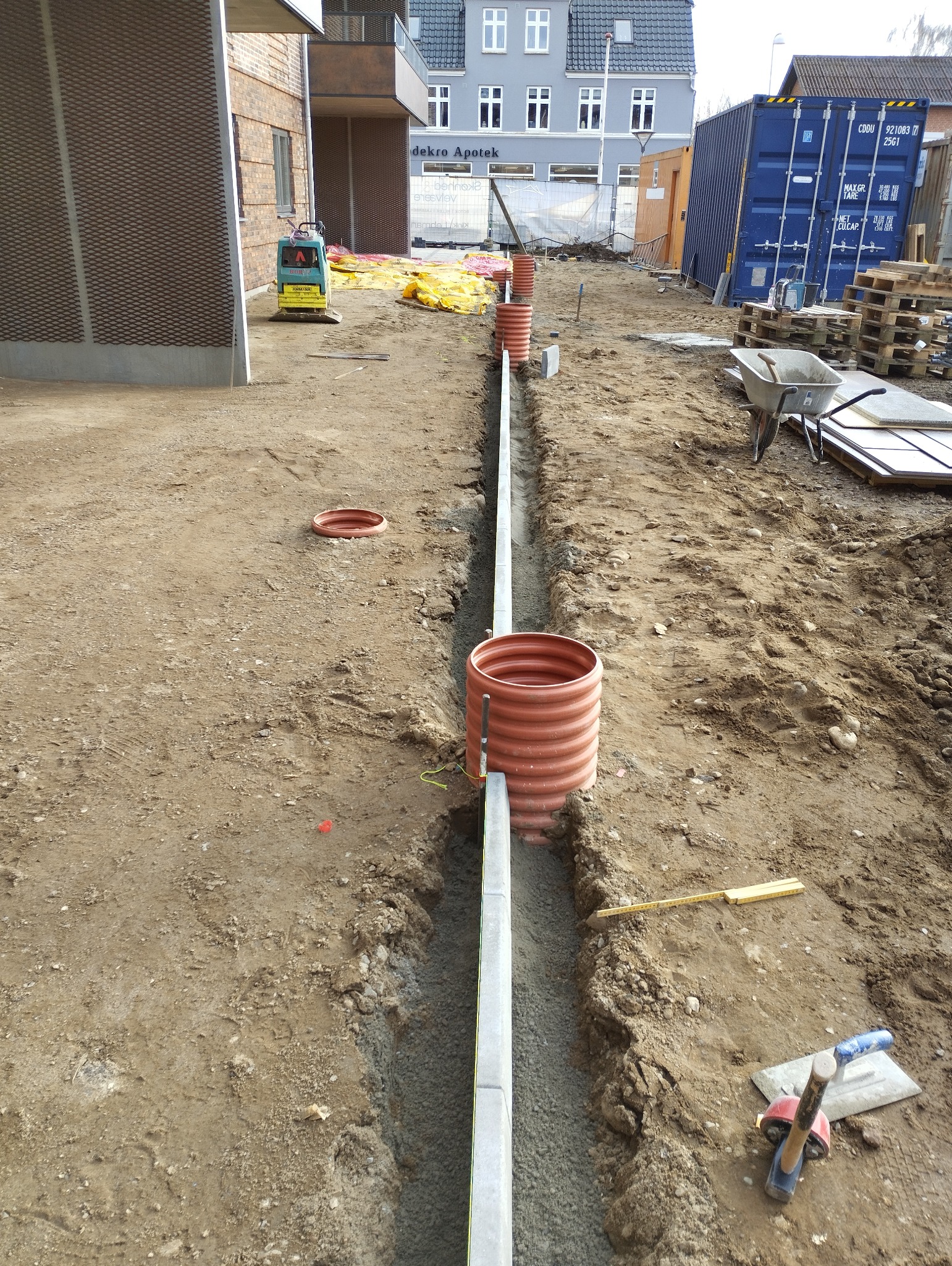 Wykop z ułożonymi obrzeżami i rurami kanalizacyjnymi na placu budowy. Widoczne narzędzia i maszyna budowlana w tle. Obrzeża betonowe, rury z tworzywa sztucznego.