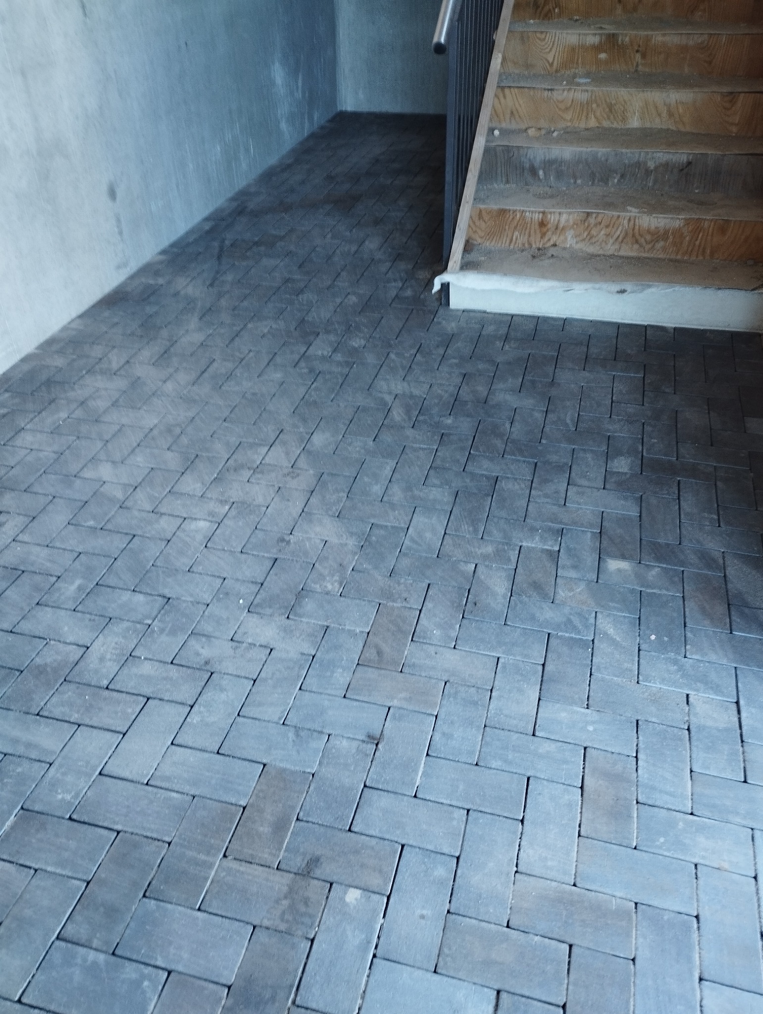 Ułożona, szara kostka brukowa w jodełkę na podłodze wewnątrz budynku, widoczne schody drewniane z metalową barierką. Surowy beton na ścianie w tle.