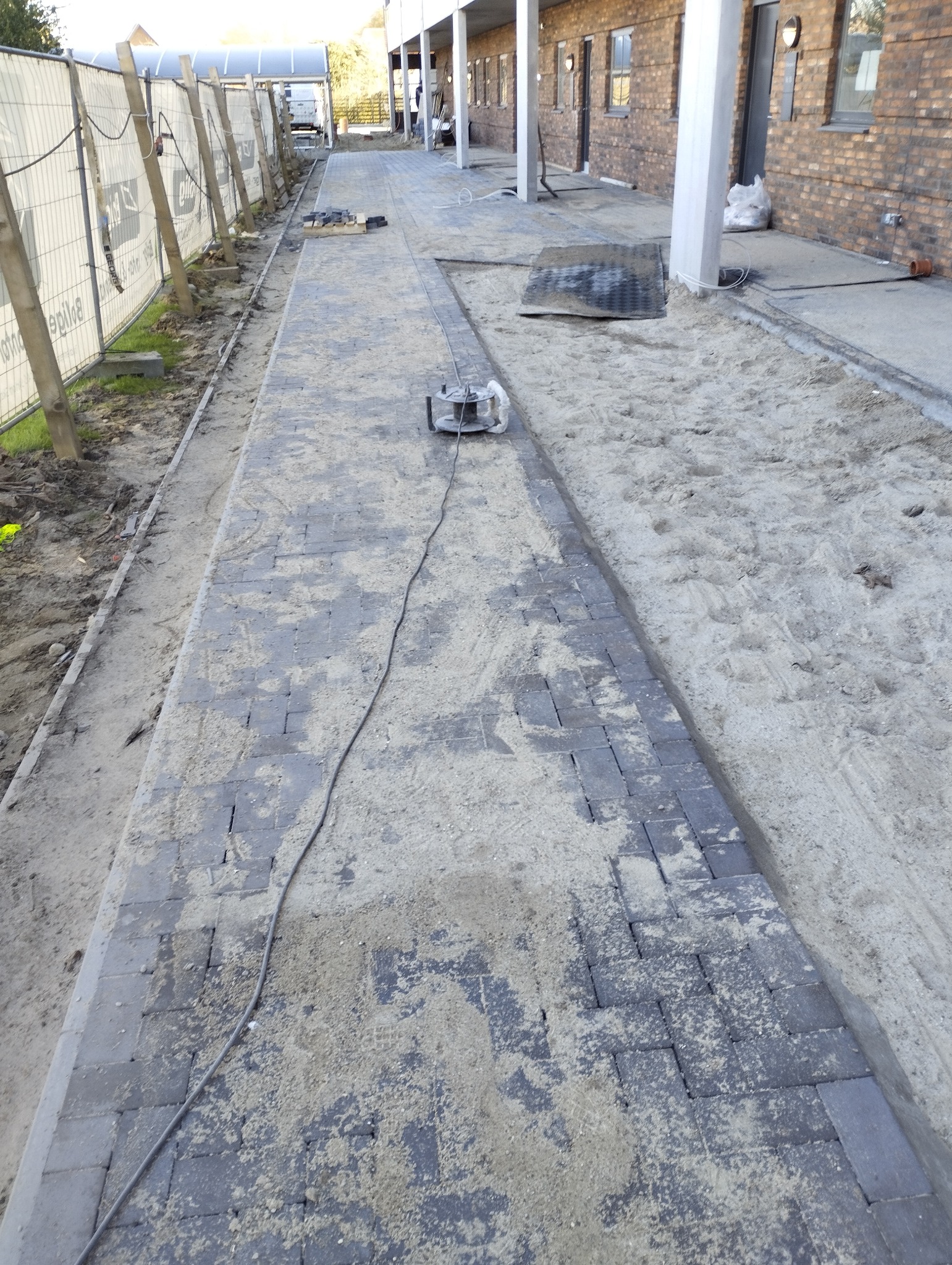 Układanie kostki brukowej na posesji, widoczny fragment ułożonej nawierzchni, piasek i narzędzie do zagęszczania. Prace brukarskie w toku, teren budowy zabezpieczony siatką.