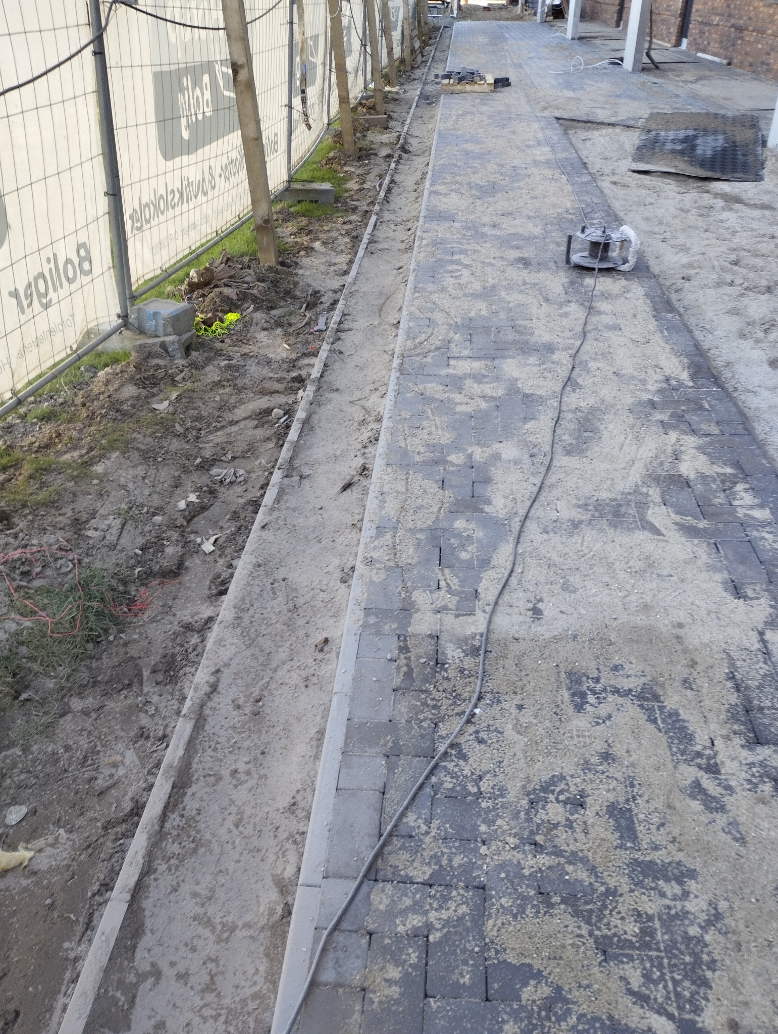 Układanie kostki brukowej na posesji, widoczne narzędzie do cięcia. Częściowo ułożona nawierzchnia z szarej kostki. Prace w toku, ogrodzenie z siatki w tle.