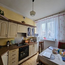 Gryfny porządek - Jasna kuchnia z drewnianymi meblami i oknem z firankami. Na stole leżą książki i okulary. Widoczny fragment podłogi z wzorem parkietu. Całość w ciepłej tonacji.