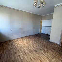 Gryfny porządek - Pusty pokój z brązowymi panelami podłogowymi i beżowymi ścianami. Widoczny fragment ściany z tapetą w tle. Na suficie wisi lampa z kilkoma szklanymi kloszami.