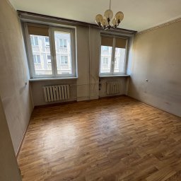 Gryfny porządek - Pusty pokój z parkietem i dwoma oknami z widokiem na budynek. Na suficie wisi lampa. Ściany w odcieniach beżu, widoczne grzejniki pod oknami.