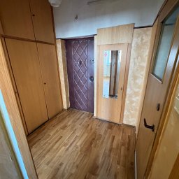 Gryfny porządek - Korytarz z brązowymi panelami podłogowymi, szafą w zabudowie, drzwiami wejściowymi z wzorem i drzwiami z lustrem. Widok na wnętrze mieszkania.