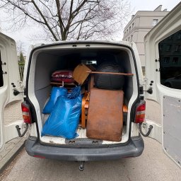 Gryfny porządek - Wnętrze białego busa wypełnione meblami i workami, widoczne otwarte tylne drzwi, drzewo w tle. Transport rzeczy podczas przeprowadzki lub opróżniania mieszkania.