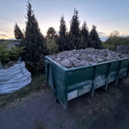 Gryfny porządek - Kontener i worek z gruzem na tle zieleni i zachodzącego słońca. Widoczne drzewa iglaste i budynek w oddali. Ujęcie z dołu, podkreślające objętość odpadów.