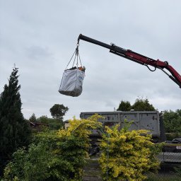 Gryfny porządek - Kran podnosi duży worek z odpadami ogrodowymi nad zarośniętym ogrodem. W tle widoczny kontener na śmieci i pochmurne niebo. Selektywna zbiórka odpadów.