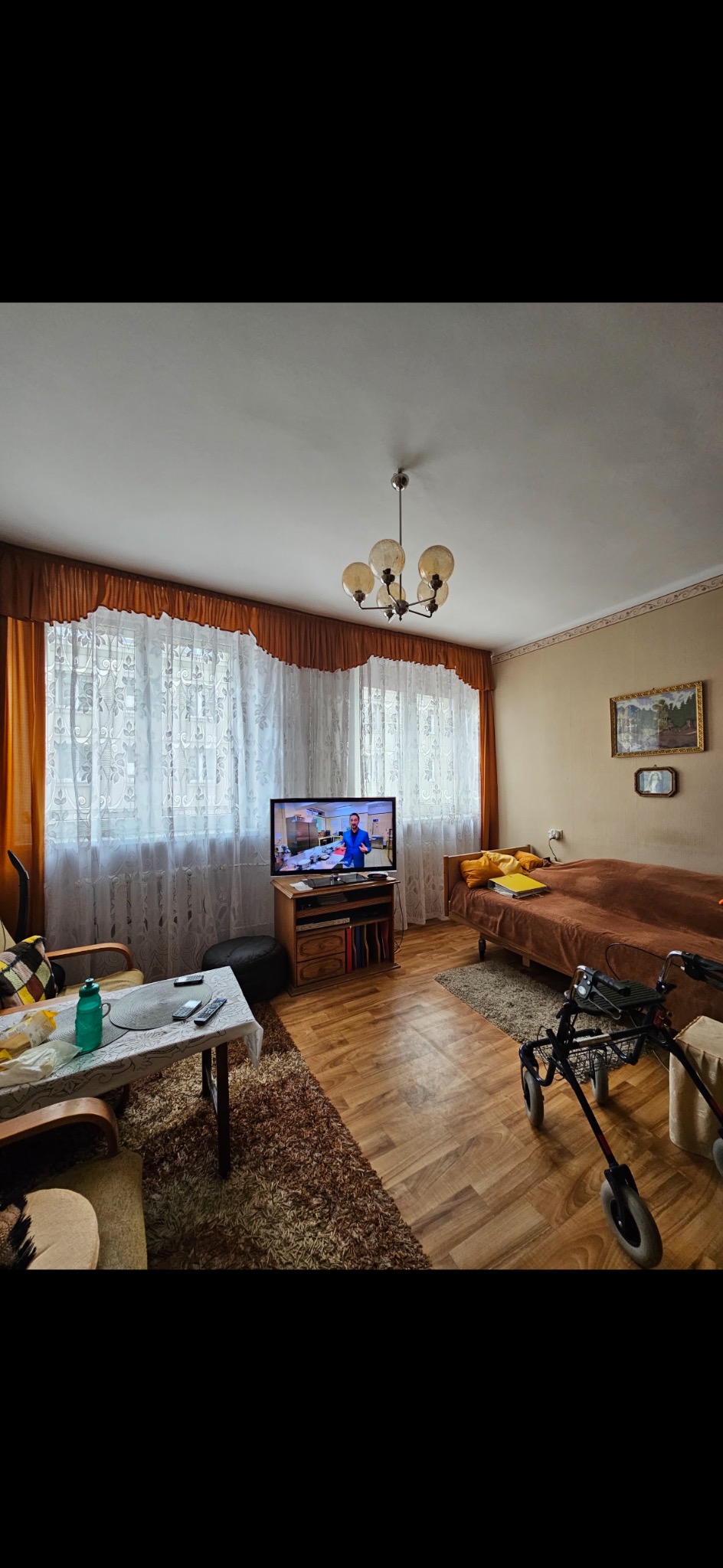 Pokój dzienny z meblami w stylu retro: brązowa kanapa, stolik z pilotami, telewizor. Na podłodze dywan shaggy. W tle okno z firankami i zasłonami.