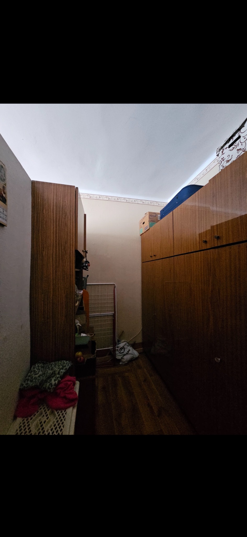 Wnętrze zagraconego pokoju z drewnianymi szafami, suszarką na pranie i workiem na podłodze. Widok na ścianę z kalendarzem. Przygotowanie do opróżniania.