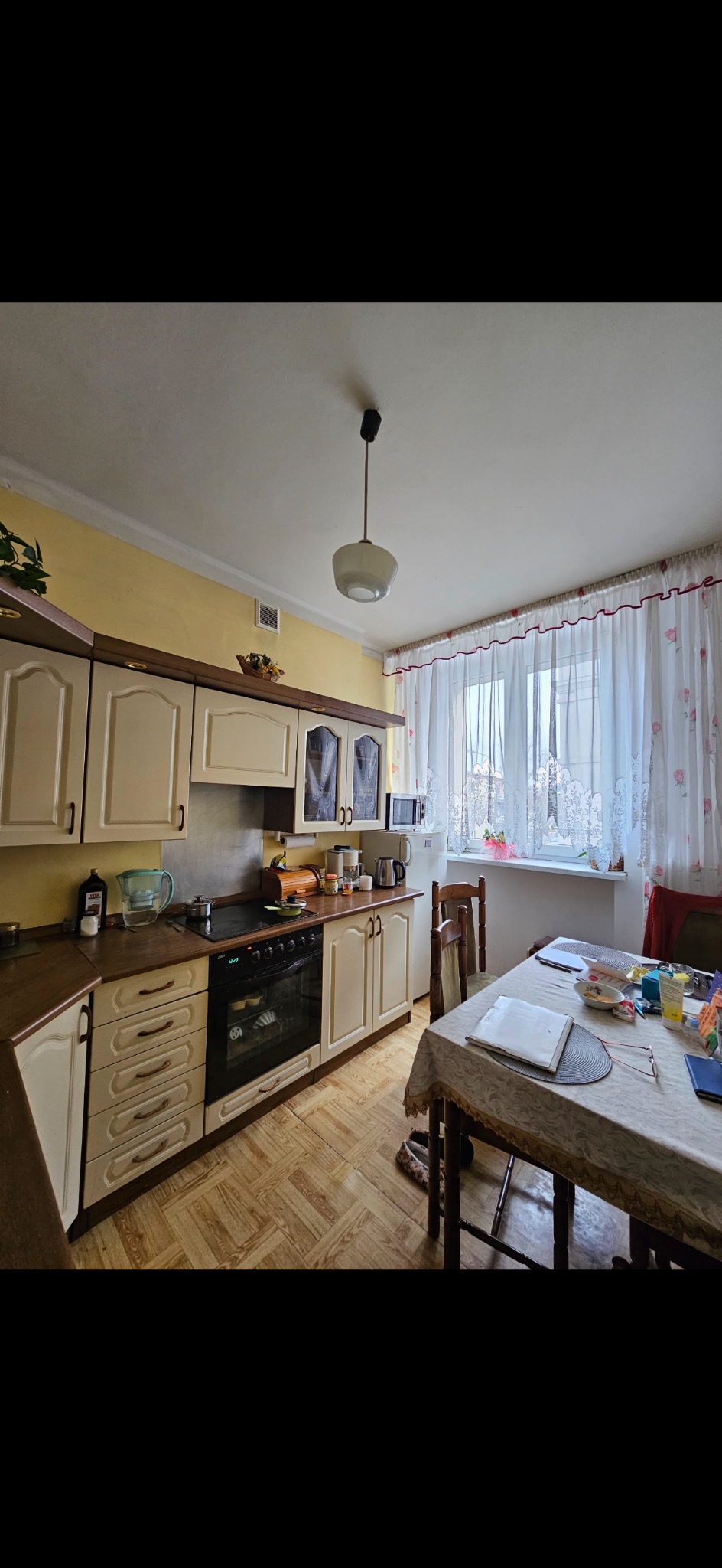 Jasna kuchnia z drewnianymi meblami i oknem z firankami. Na stole leżą książki i okulary. Widoczny fragment podłogi z wzorem parkietu. Całość w ciepłej tonacji.