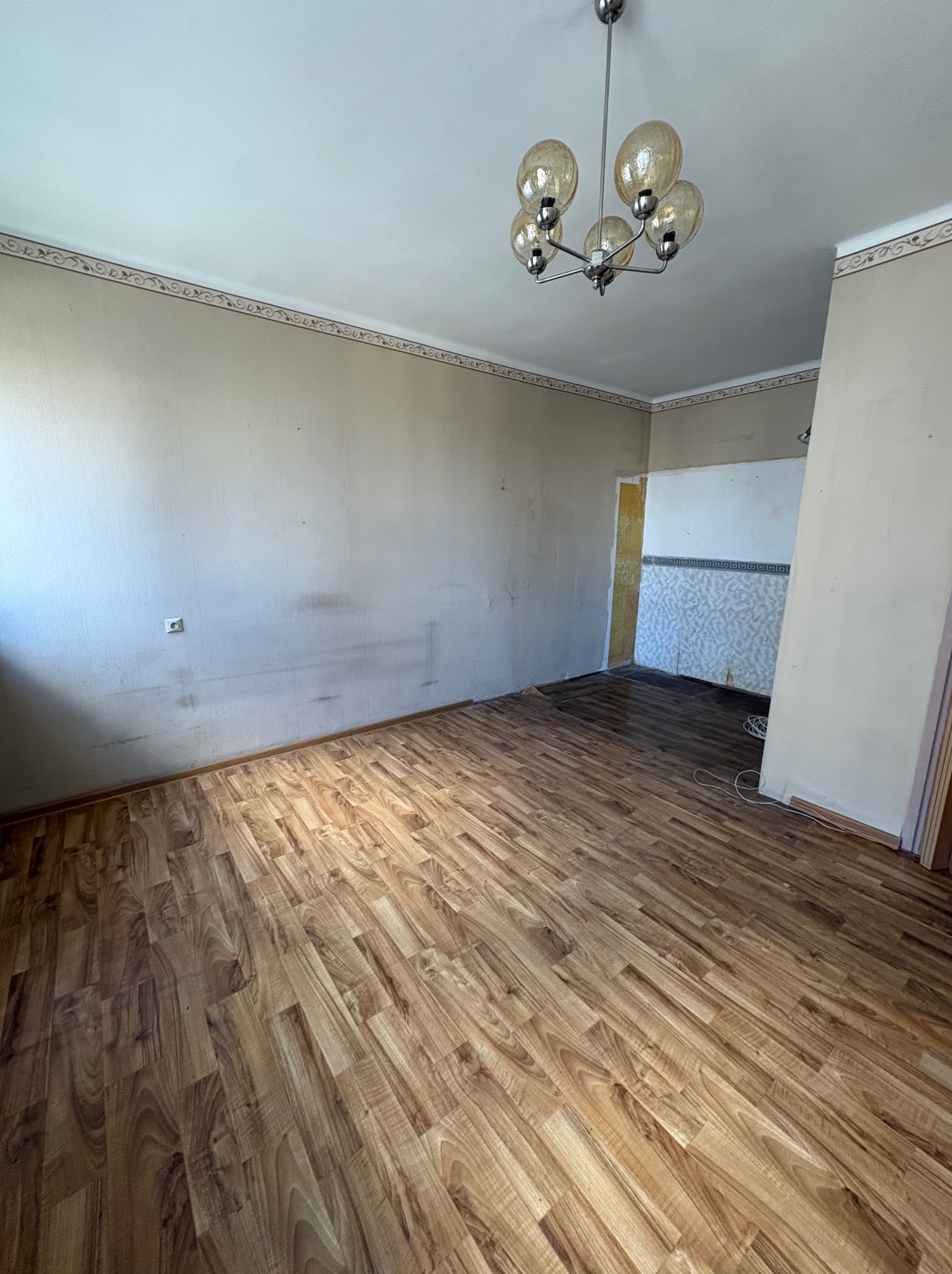 Pusty pokój z brązowymi panelami podłogowymi i beżowymi ścianami. Widoczny fragment ściany z tapetą w tle. Na suficie wisi lampa z kilkoma szklanymi kloszami.