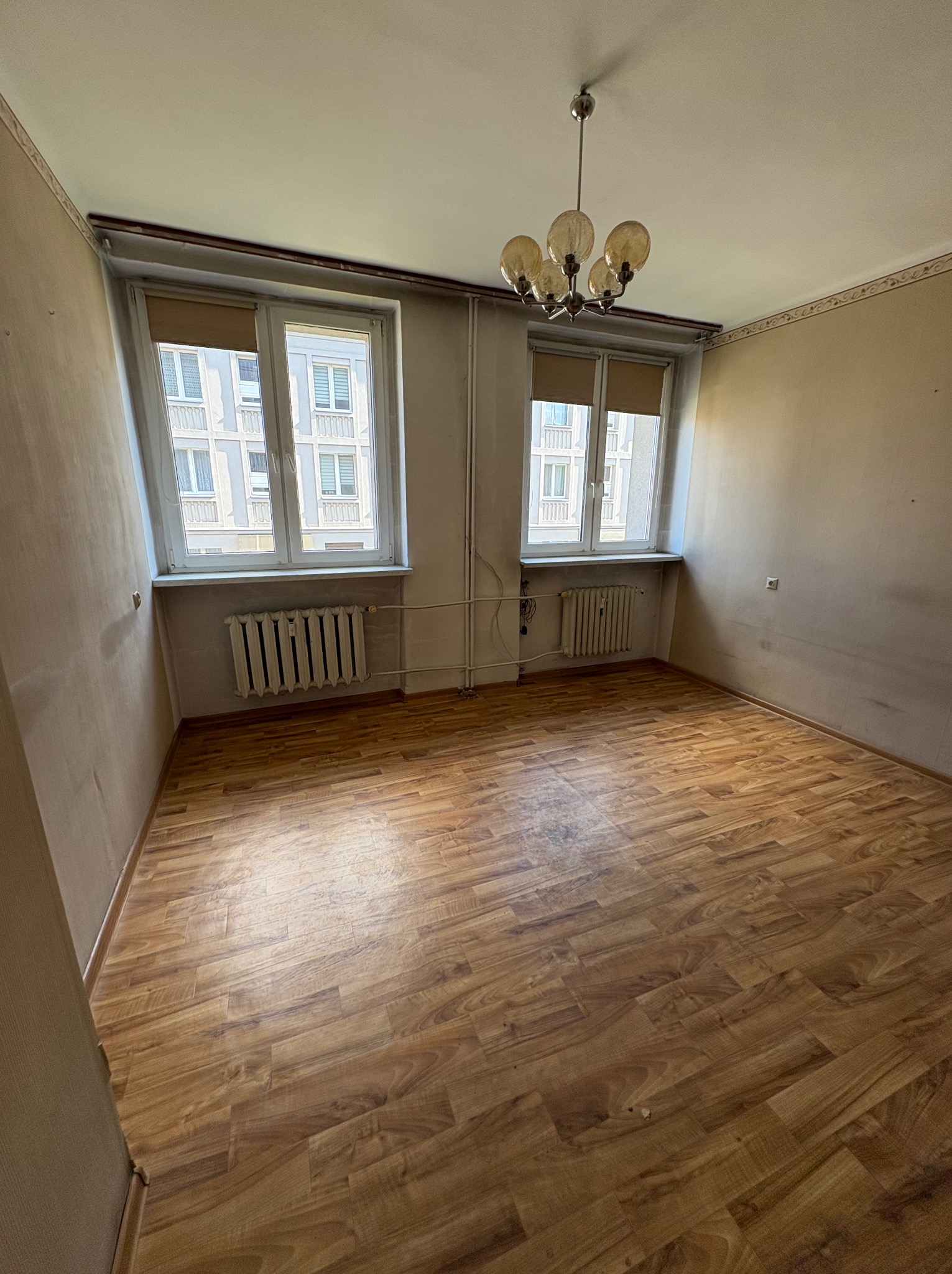 Pusty pokój z parkietem i dwoma oknami z widokiem na budynek. Na suficie wisi lampa. Ściany w odcieniach beżu, widoczne grzejniki pod oknami.