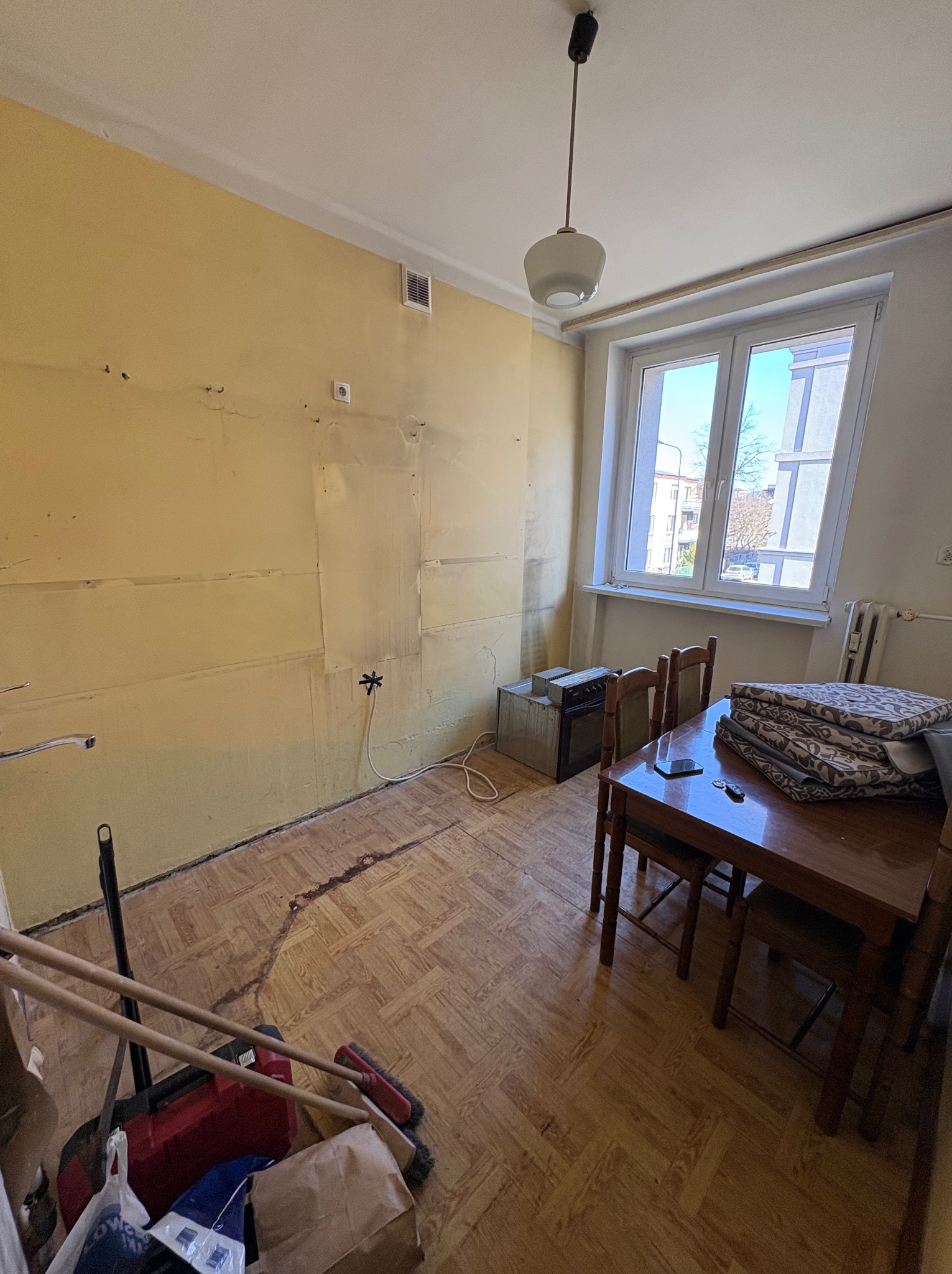 Pokój przed remontem: żółte ściany z resztkami tapet, parkiet, stół z krzesłami, sprzęty. Widoczne ślady demontażu i przygotowania do prac remontowych.