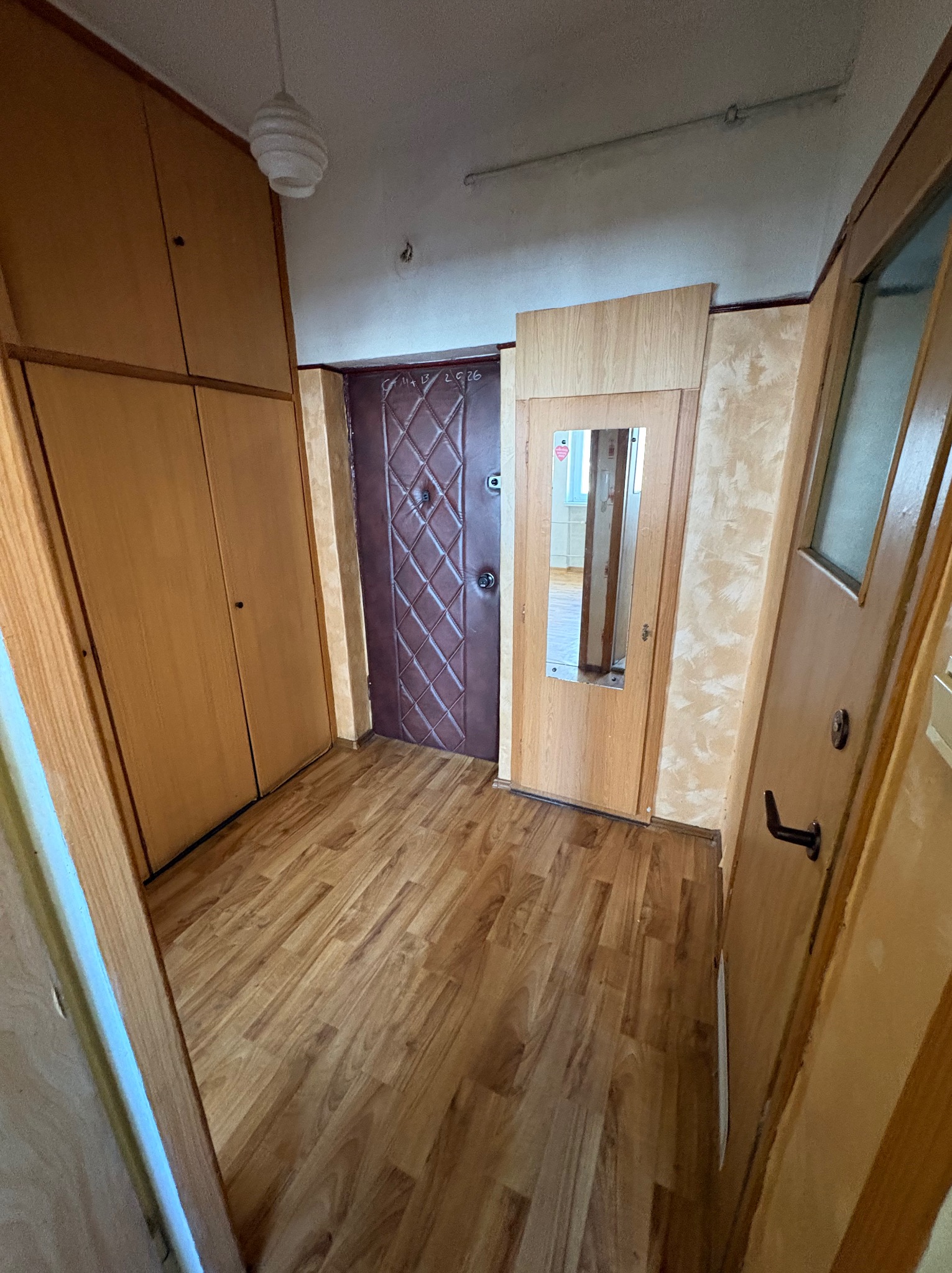 Korytarz z brązowymi panelami podłogowymi, szafą w zabudowie, drzwiami wejściowymi z wzorem i drzwiami z lustrem. Widok na wnętrze mieszkania.