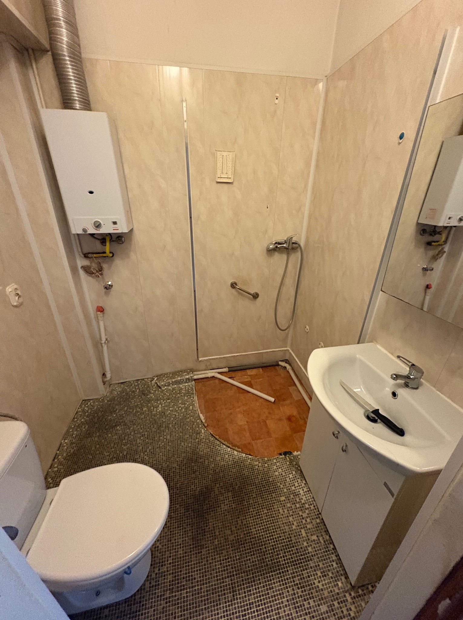 Łazienka w trakcie demontażu: częściowo zerwane płytki podłogowe, odłączony bojler, umywalka z nożem. Widoczne ślady po demontażu.