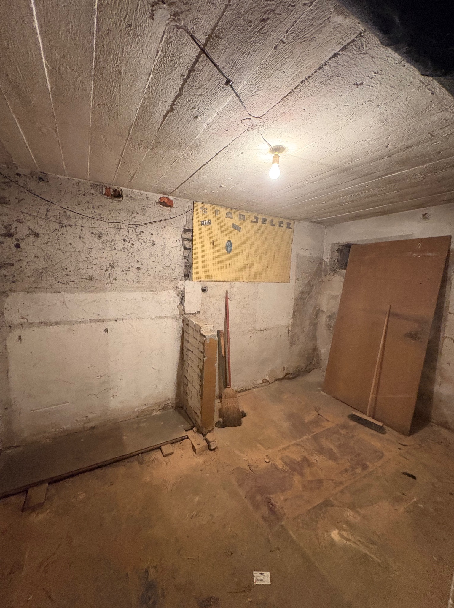 Piwnica w trakcie opróżniania: widoczne ściany z resztkami farby, podłoga pokryta kurzem i gruzem, oraz narzędzia jak miotła i płyta. Żarówka oświetla surowe wnętrze.