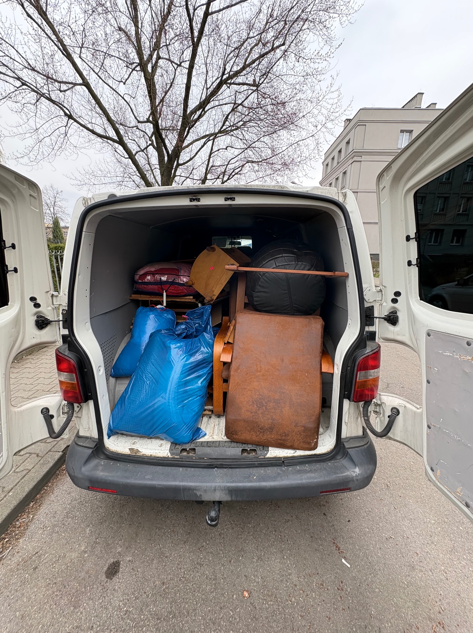 Wnętrze białego busa wypełnione meblami i workami, widoczne otwarte tylne drzwi, drzewo w tle. Transport rzeczy podczas przeprowadzki lub opróżniania mieszkania.