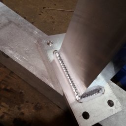 Rafał Małolepszy - Precyzyjne spawy na aluminiowej konstrukcji, widoczne regularne spoiny. Detal złącza kątowego z otworami montażowymi, surowe wykończenie metalu.