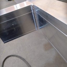 Rafał Małolepszy - Szczegółowe ujęcie spawu łączącego dwa aluminiowe profile pod kątem prostym. Widoczna precyzja wykonania i gładka powierzchnia metalu. W tle szary beton.