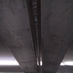 Rafał Małolepszy - Metalowa konstrukcja z widocznym spoiwem spawalniczym, ukazana z perspektywy od dołu, z elementami oświetlenia w tle. Szara kolorystyka i surowy wygląd metalu.