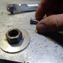Rafał Małolepszy - Detal z warsztatu: nakrętka przyspawana do podkładki, element metalowy z widocznym spawem i dłoń trzymająca mały, metalowy cylinder na tle metalowego blatu.
