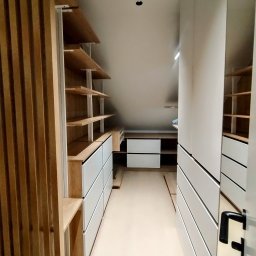 Piotr Białousz Woodshore - Funkcjonalna garderoba na poddaszu ze skośnym sufitem. Jasne drewno i białe fronty szuflad. Otwarta przestrzeń z półkami i szufladami, minimalistyczny design.