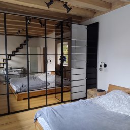 Piotr Białousz Woodshore - Nowoczesna sypialnia z szafą wnękową z czarnymi ramami, odbijającą schody i łóżko. Widoczne drewniane belki sufitowe i oświetlenie punktowe.