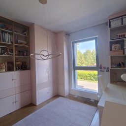 Piotr Białousz Woodshore - Jasny pokój z zabudową meblową: przeszklona biblioteczka, szafa z frezowanymi frontami i regał z dekoracjami. Okno z widokiem na zieleń. Całość w pastelowej kolorystyce.