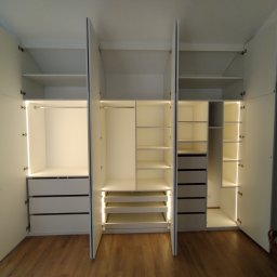 Piotr Białousz Woodshore - Otwarta, biała szafa wnękowa z oświetleniem LED, ukazująca półki, szuflady i drążki na ubrania. Widoczny parkiet na podłodze i fragment sypialni w tle.