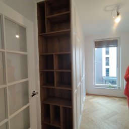 Piotr Białousz Woodshore - Wnętrze z drewnianą szafą wnękową z otwartymi półkami, przylegającą do białej szafy zamykanej, na parkiecie w jodełkę, z widokiem na okno.