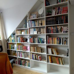 Piotr Białousz Woodshore - Biała, zabudowana biblioteczka na książki pod skosem dachu, wypełniona książkami, dekoracjami i pamiątkami. Widoczne drewniane półki i różnorodna kolorystyka okładek.