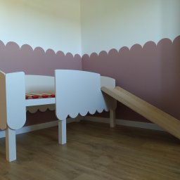 Piotr Białousz Woodshore - Białe łóżko dziecięce z drewna ze zjeżdżalnią, na podłodze panele, ściana w pastelowych kolorach. Nietypowy kształt ramy łóżka.