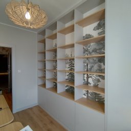 Piotr Białousz Woodshore - Biała szafa wnękowa z drewnianymi półkami i tapetą w leśny motyw. Nowoczesne oświetlenie sufitowe. Widok na wnętrze przez otwarte drzwi.