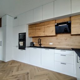 Piotr Białousz Woodshore - Nowoczesna kuchnia z białymi szafkami i drewnianymi akcentami. Czarny blat i armatura. Parkiet w jodełkę. Minimalistyczny design z wbudowanym sprzętem.