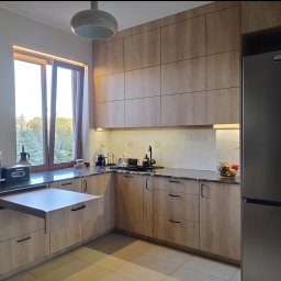 Piotr Białousz Woodshore - Jasna kuchnia z drewnianymi frontami, czarnym blatem i wysuwanym blatem roboczym. Widoczne oświetlenie podszafkowe i okno z widokiem na zieleń.