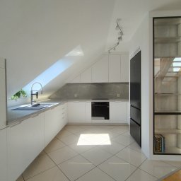 Piotr Białousz Woodshore - Jasna kuchnia na poddaszu z białymi szafkami, szarym blatem i czarną lodówką. Uwagę przykuwa przeszklona szafka z podświetleniem i książkami na półce.