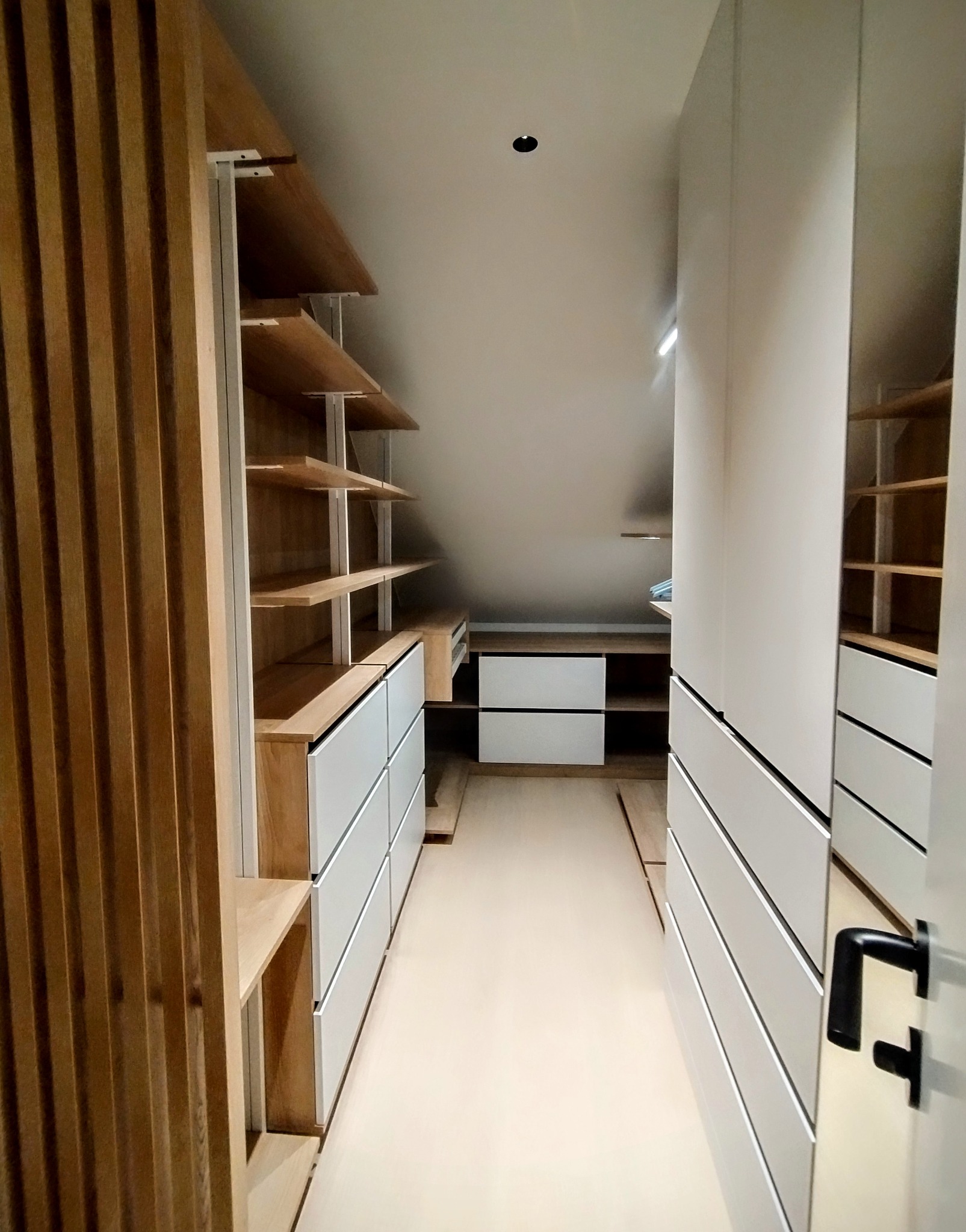 Funkcjonalna garderoba na poddaszu ze skośnym sufitem. Jasne drewno i białe fronty szuflad. Otwarta przestrzeń z półkami i szufladami, minimalistyczny design.