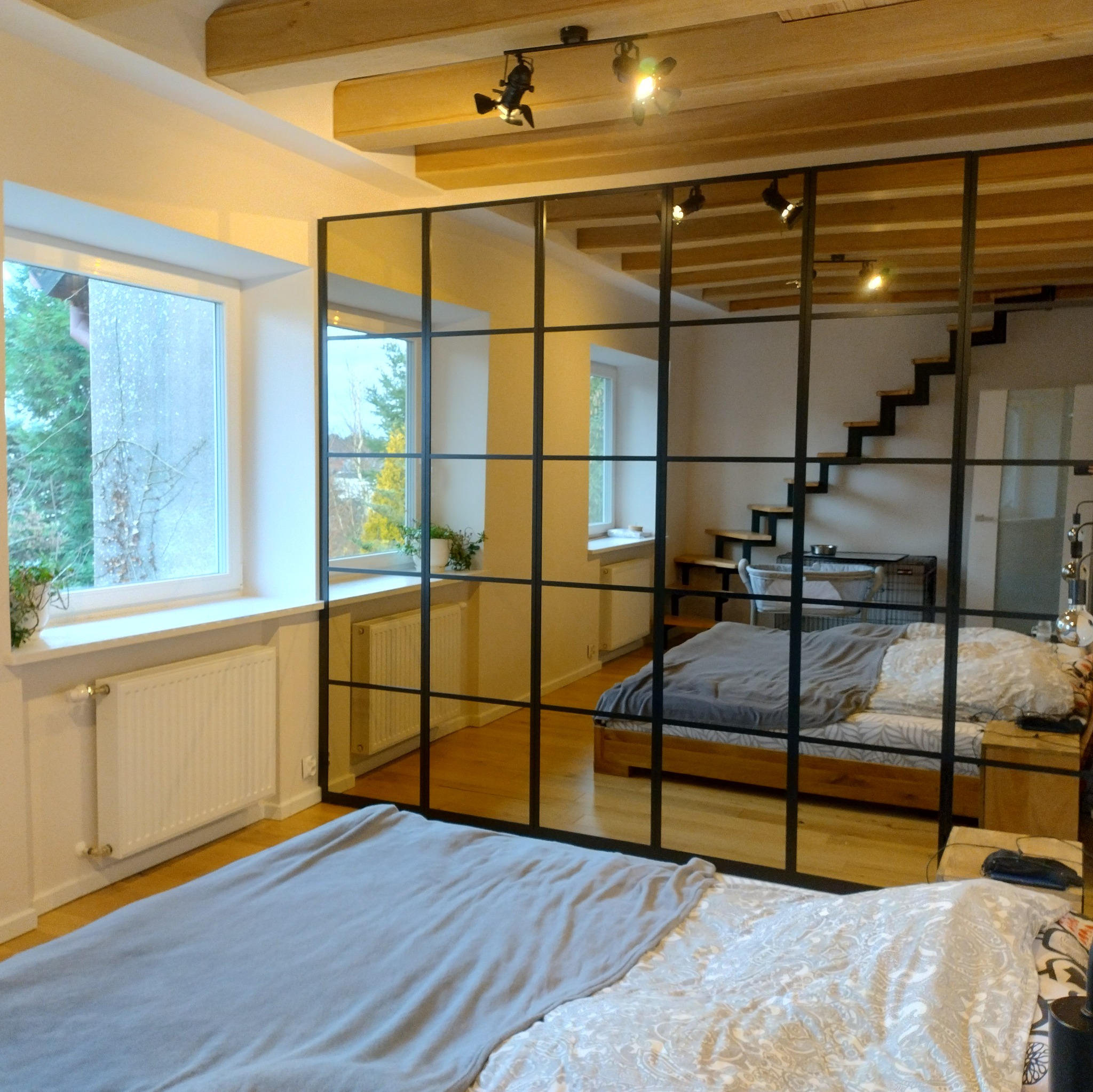 Loftowa sypialnia z drewnianym stropem i ścianką działową z czarnych profili. Widoczne dwa łóżka, schody i okno z widokiem na zieleń. Minimalistyczny design.