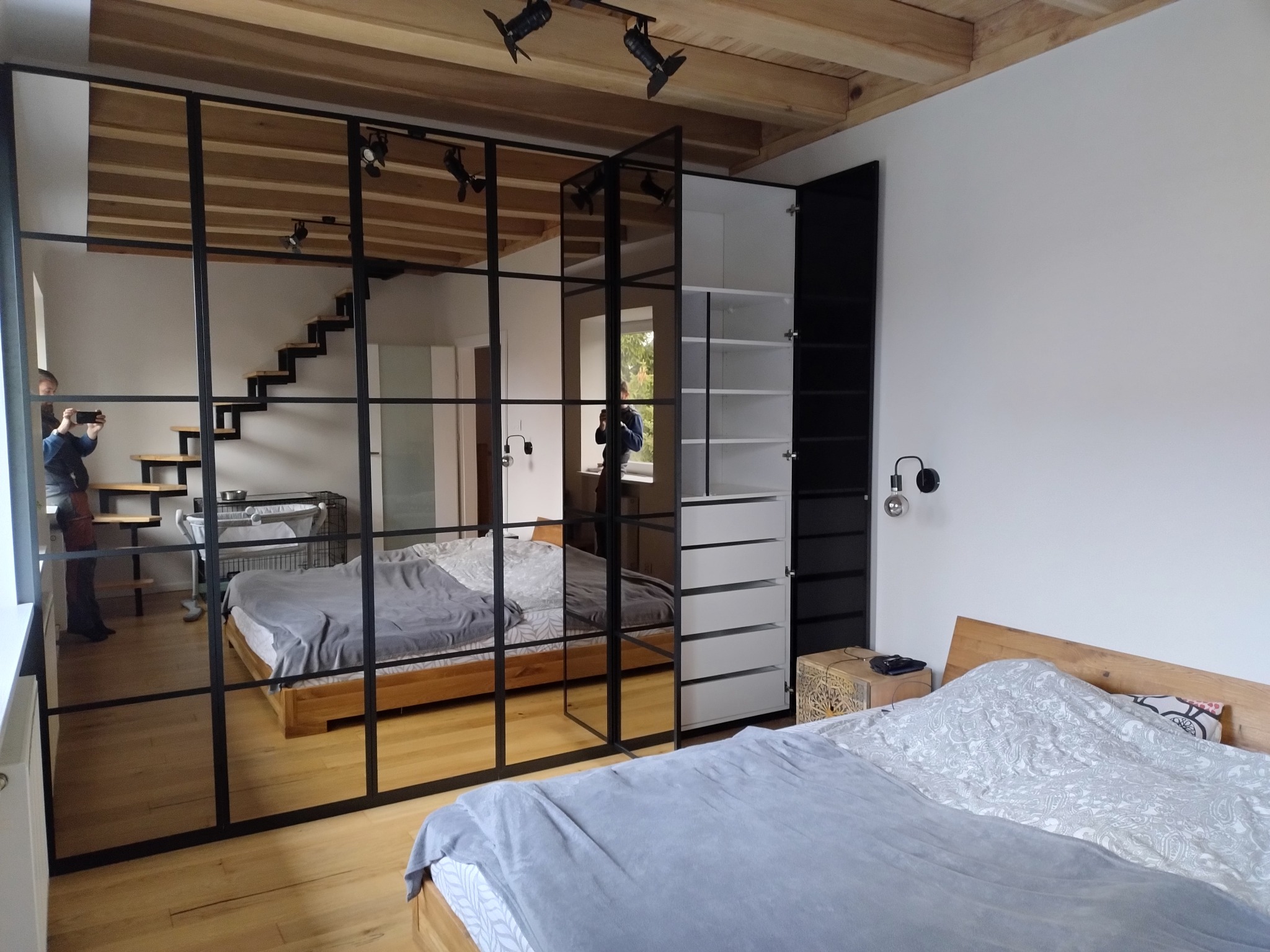 Nowoczesna sypialnia z szafą wnękową z czarnymi ramami, odbijającą schody i łóżko. Widoczne drewniane belki sufitowe i oświetlenie punktowe.